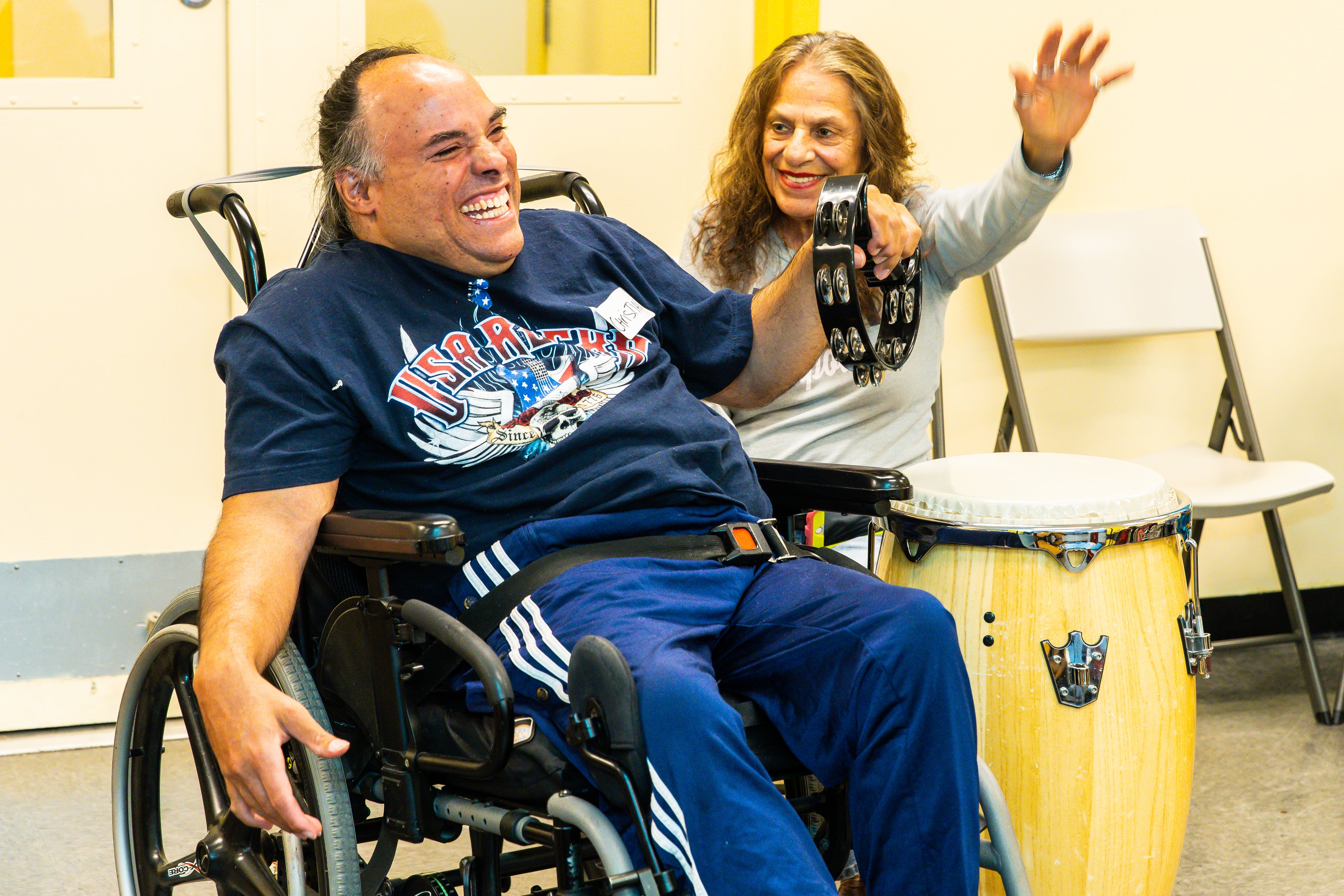 Image of a member at DMF playing the tamborine next to an older woman. He is using a wheelchair, and smiling broadly. 